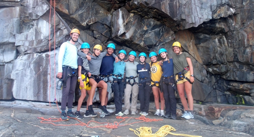 A group of people wearing safety gear stand in a line and smile for a group photo. They are in front of a rock wall.